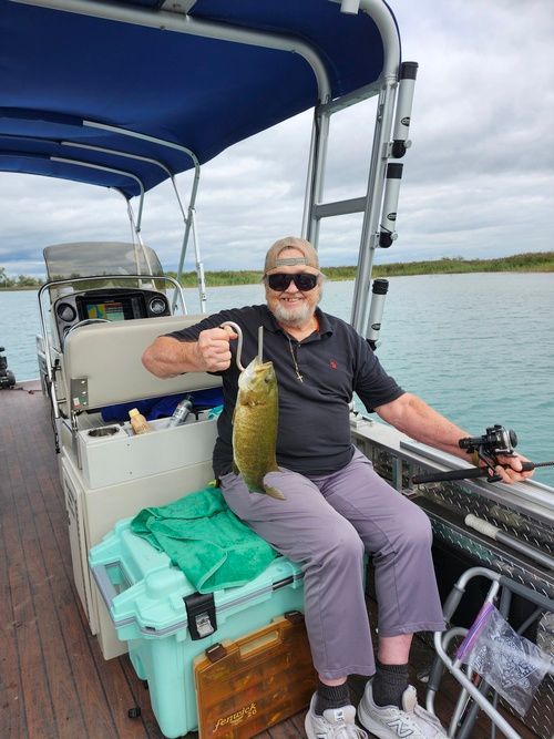 Man on a pontoon boat, holding up a fish. Cloudy sky, water in the background.