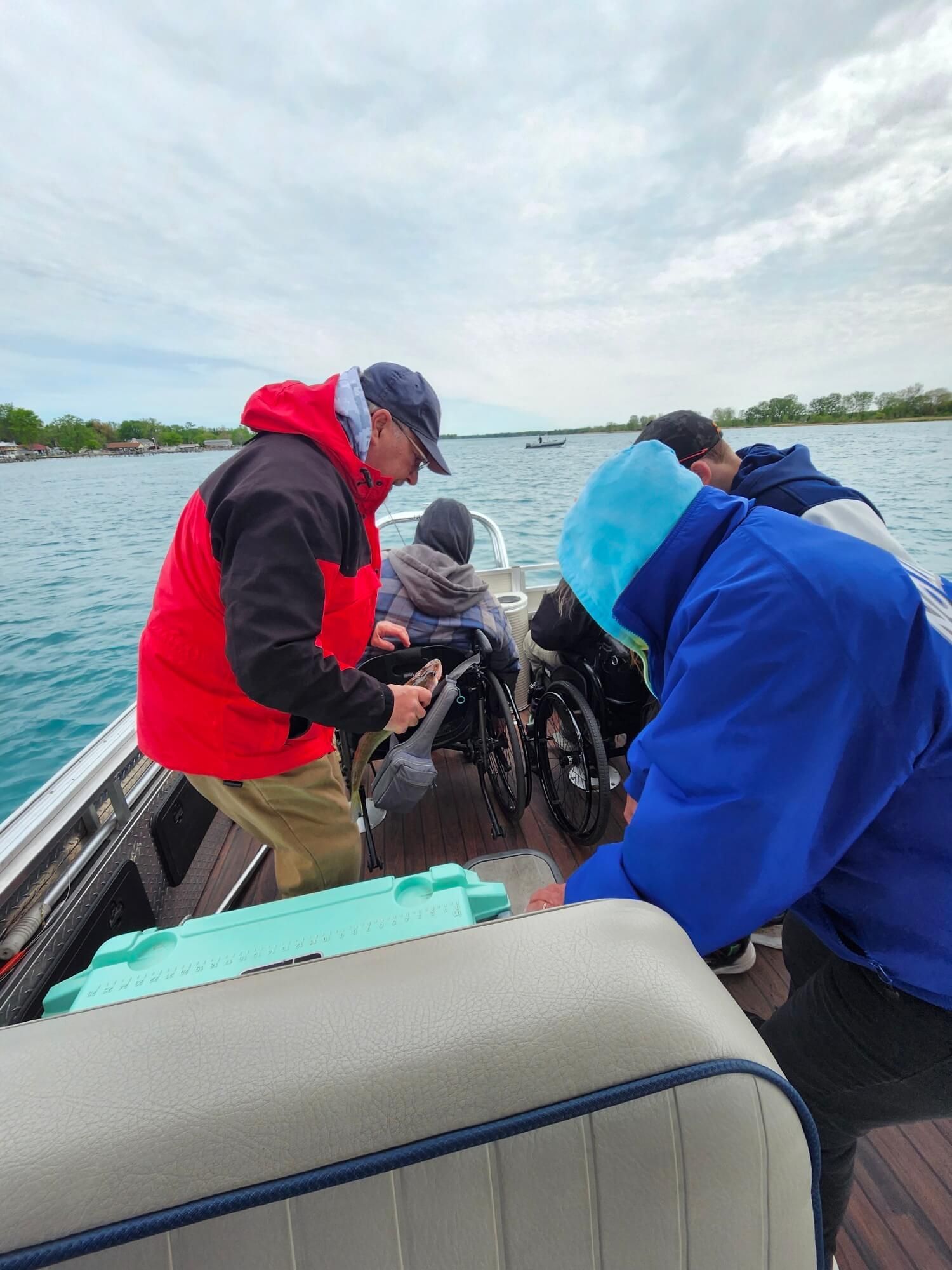 Three people in rain jackets work on a boat engine on a lake under cloudy skies