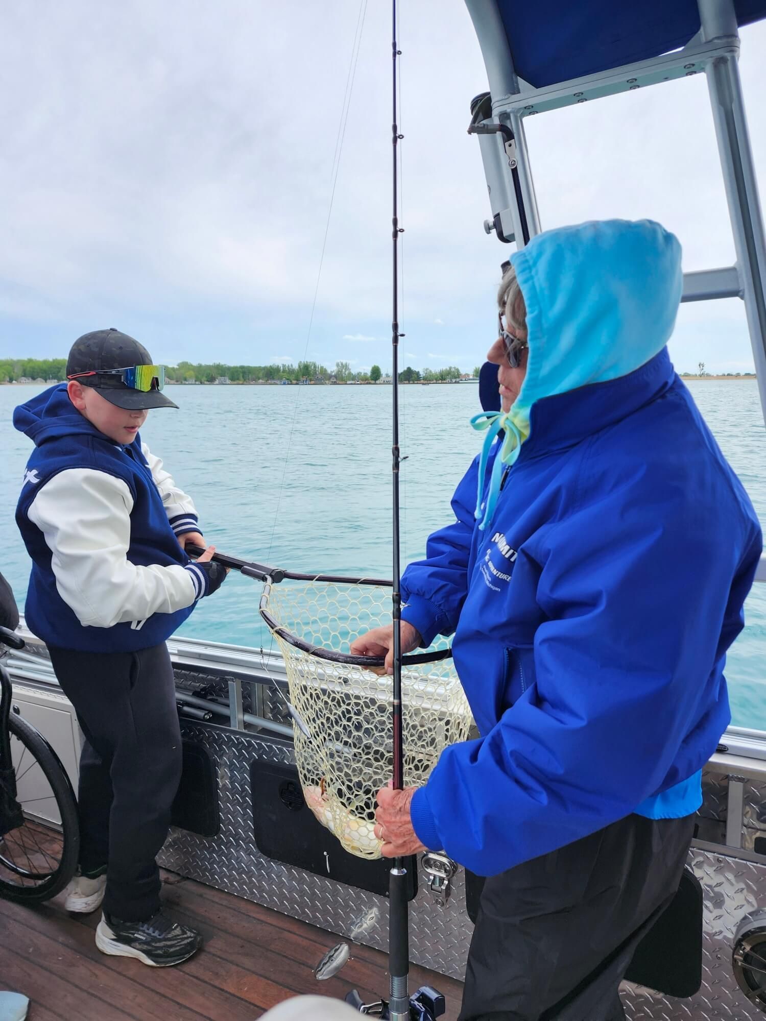 Two people on a boat deck handling a fishing net on calm water under a cloudy sky