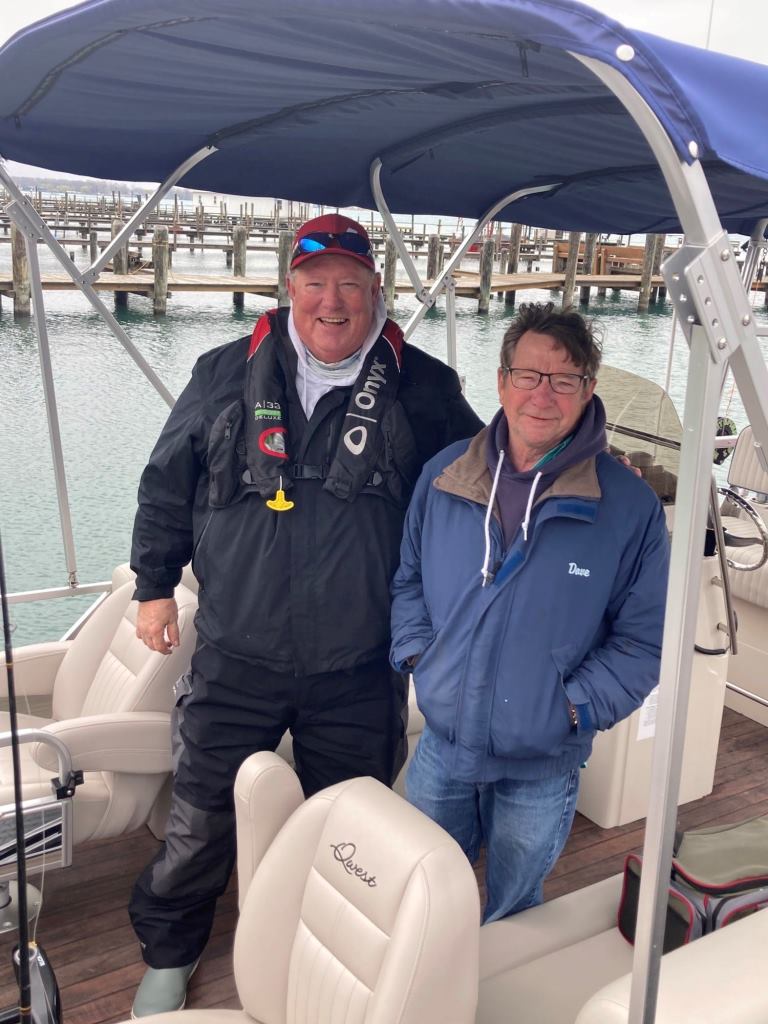 Two men standing on a pontoon boat; one in a life vest and a cap, the other in a blue jacket.