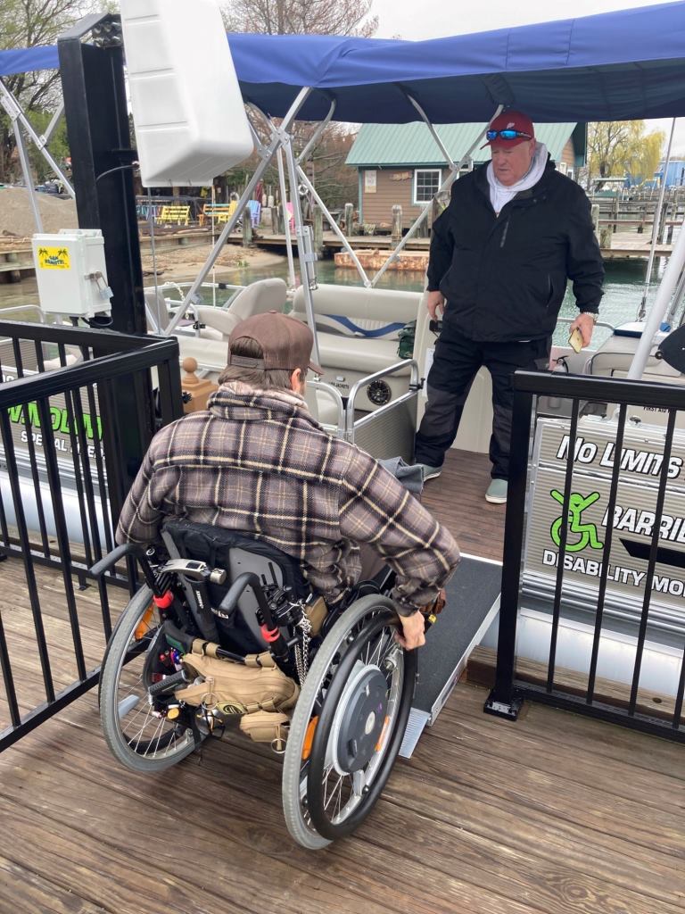 Man in wheelchair using a ramp onto a pontoon boat, assisted by another man on the boat.