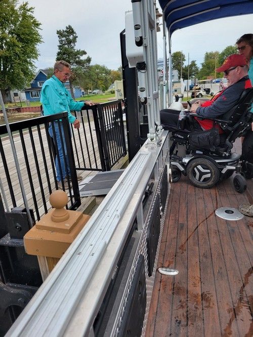 Man in wheelchair boards pontoon boat via ramp. Another man assists at dock.