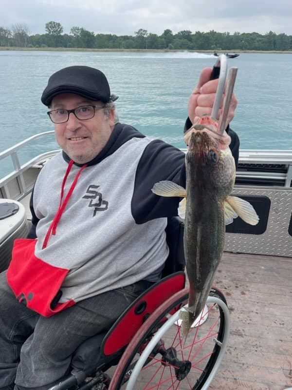 Man in wheelchair on a boat holding a fish up with a scale. He is smiling.