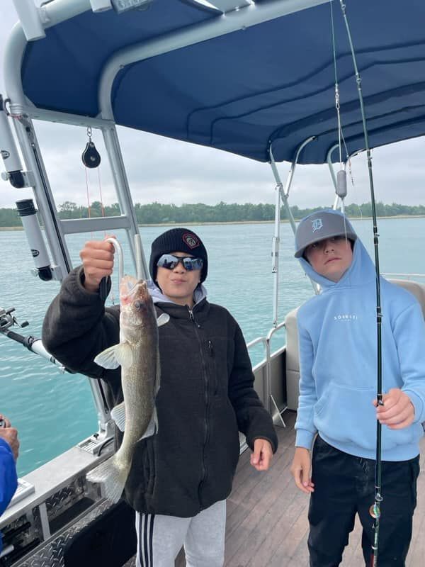 Two people on a boat, one holding a fish. Cloudy day, water visible.