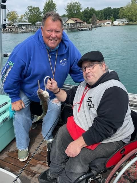 Two men on a boat; one holding up a fish on a hook. The other man is smiling. Lake in the background.