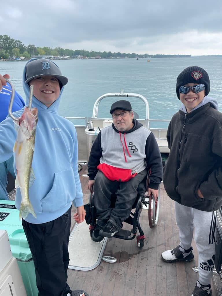 Two boys and a person in a wheelchair on a boat. One boy holds a fish. Lake in the background.