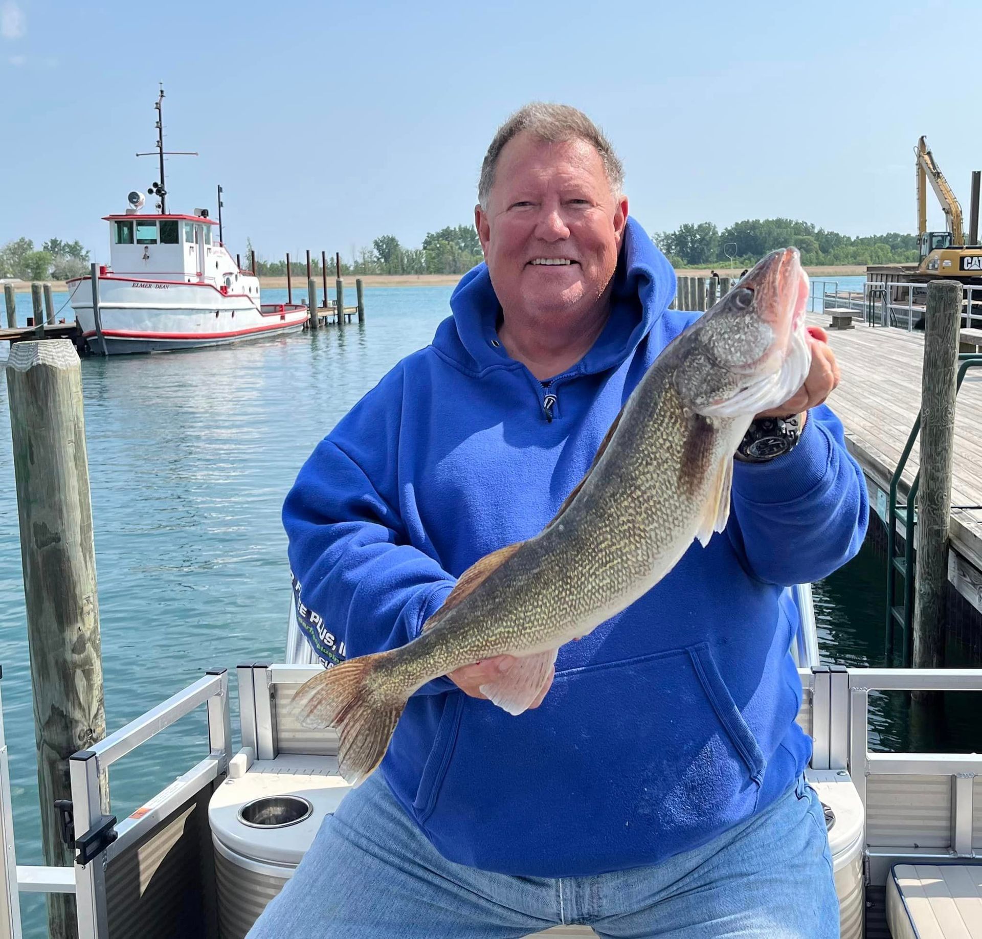 Man in blue hoodie holding a fish, near a dock with a boat.