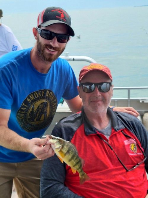 Man holding a fish, smiling with another man. Outdoors; both wearing sunglasses.