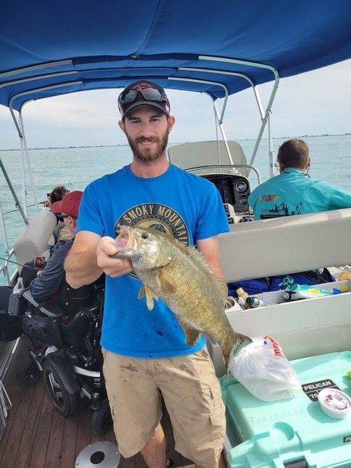 Man on a boat holding a smallmouth bass; light blue water, cloudy sky.