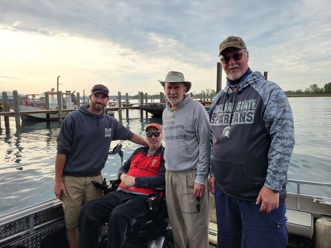 Four men on a boat near a dock. One man is in a wheelchair. Cloudy sky, water in the background.