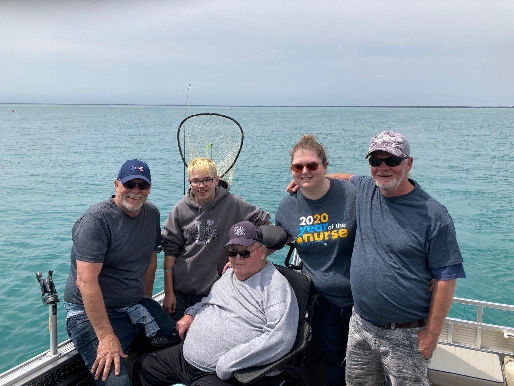 Five people on a boat, one in a wheelchair, on blue water. Cloudy sky.