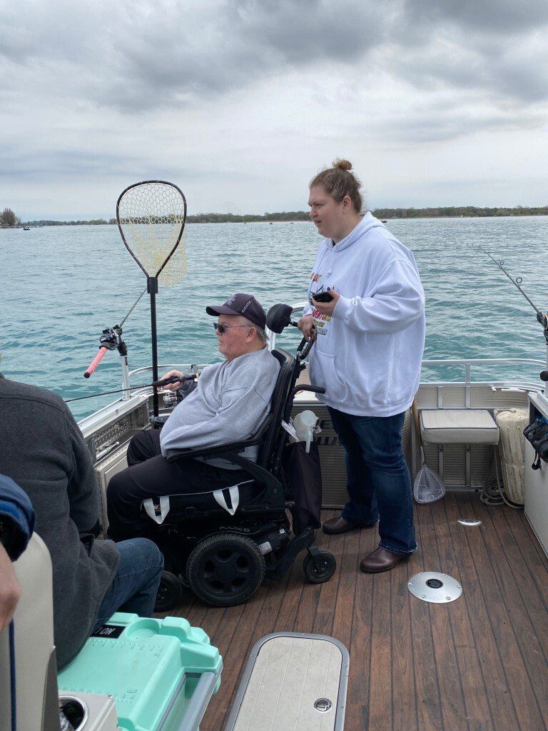 Person in wheelchair fishing on a boat, assisted by another person. Lake with overcast sky.