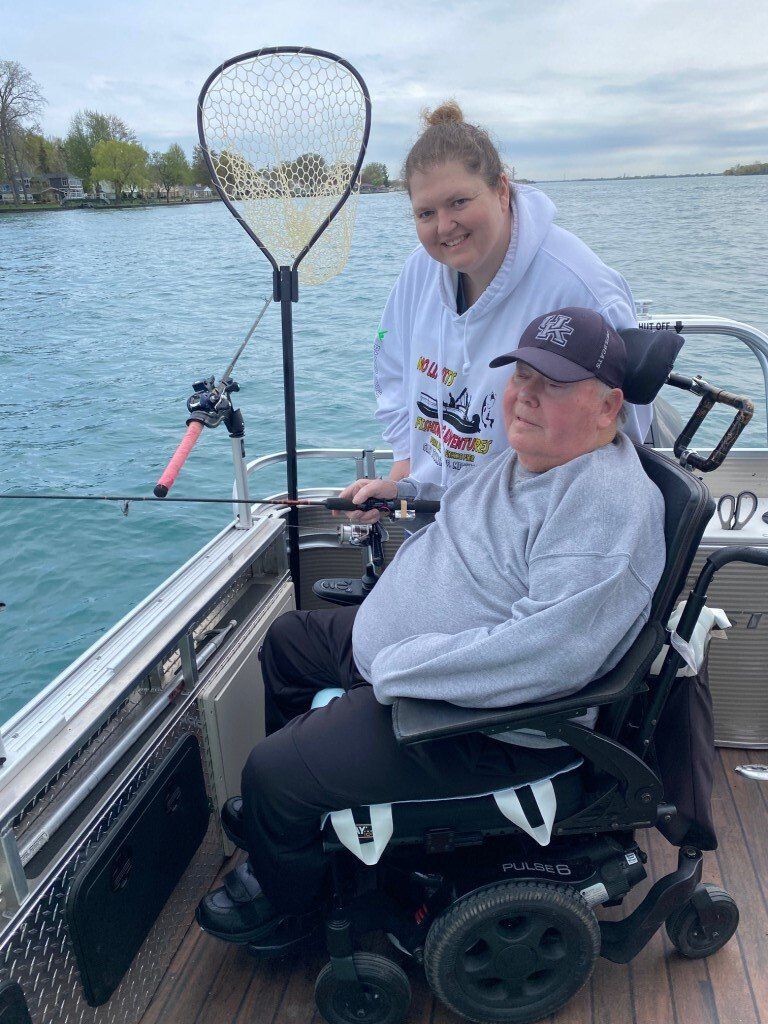 Woman helping a person in a wheelchair fish on a boat. The man is holding a fishing rod. Water and land visible in background.