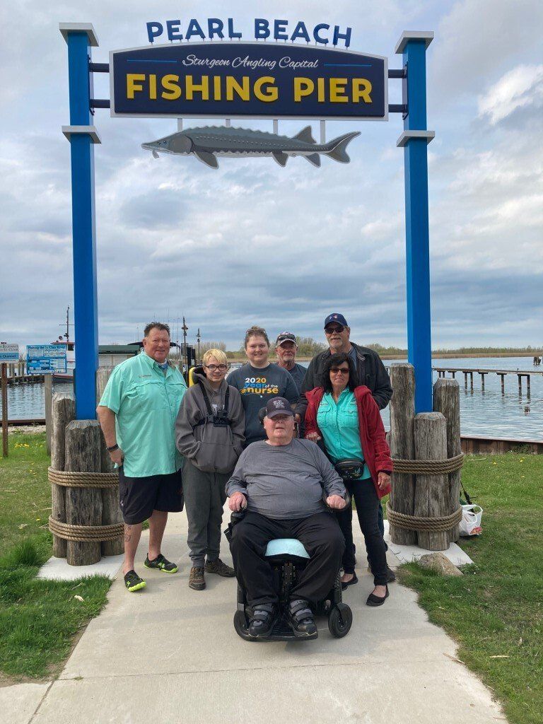 Group poses at Pearl Beach Fishing Pier sign. People, one in wheelchair, stand on a walkway near the pier.