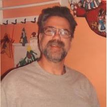Man with glasses and a beard smiles, in front of a colorful wall with hanging decorations.