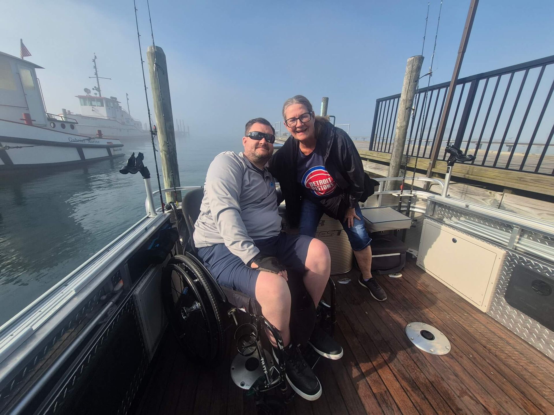 Two people sitting on a wooden dock beside a boat, smiling in a sunny marina.