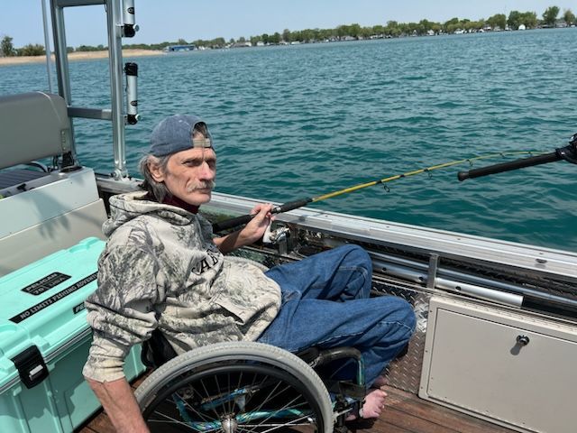 Man in a wheelchair fishing from a boat on a lake, holding a fishing rod.