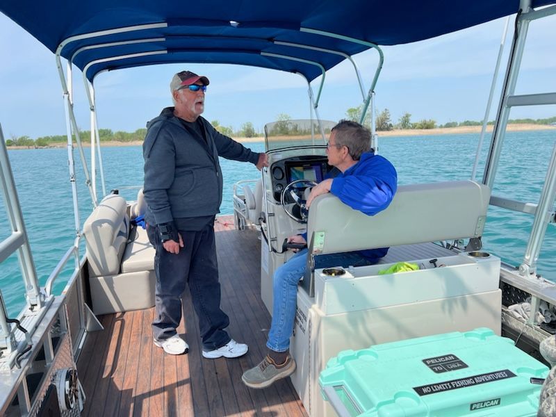 Two people on a pontoon boat on water. One stands, the other sits at the helm. Sunny day.