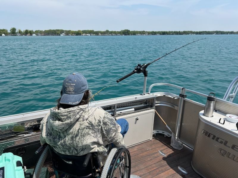 Person in wheelchair fishing from a pontoon boat on a lake; sunny day.