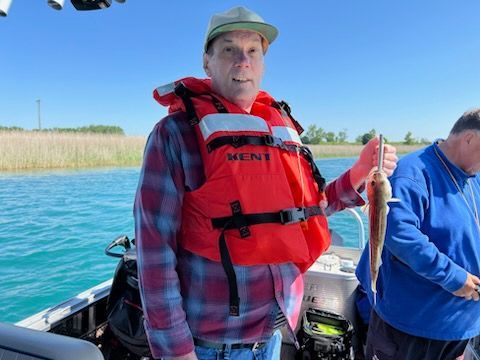 Person on a boat holding a small fish, wearing a life vest and plaid shirt, blue water in the background.