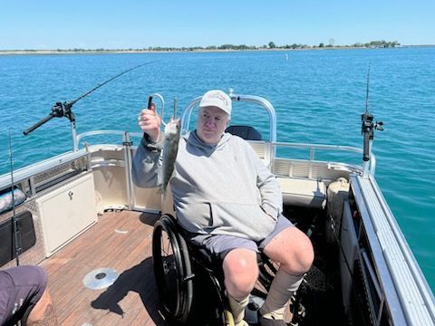 Man in wheelchair fishing from a boat, holding up a fish. Blue water and sky.
