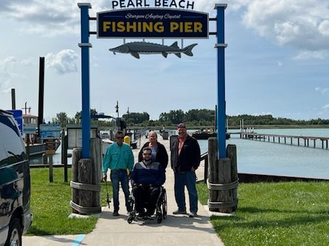 Four people by a fishing pier sign; one in a wheelchair. Sunny day, pier extends into water.