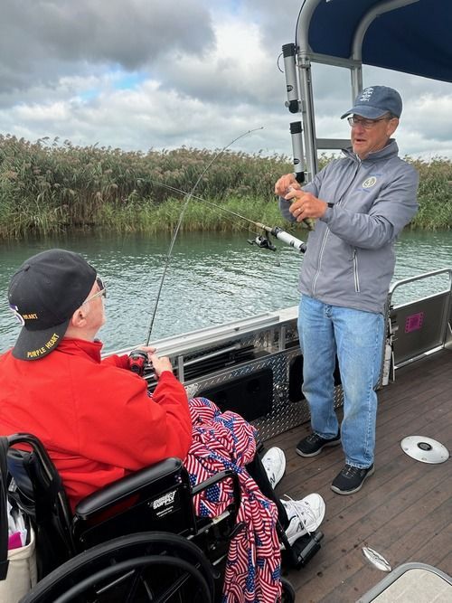 Man in wheelchair fishing with assistance on a boat. Another man helps, pointing at a fishing rod. Water and reeds in background.