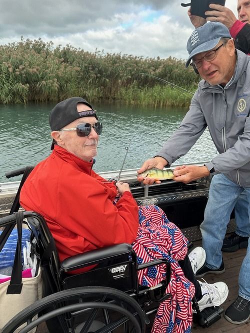 Man in wheelchair on a boat holding a fishing rod, another man showing him a caught fish.
