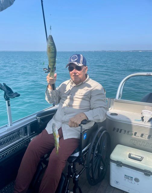 Man in wheelchair on a boat holding a caught fish, fishing rod above. Overcast day.