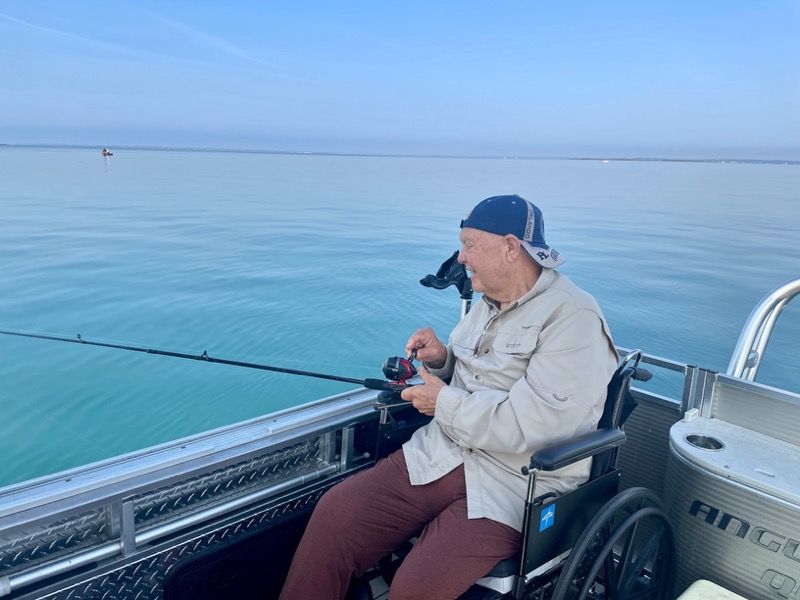 Man in wheelchair fishing on a boat in calm water, wearing a hat, shirt, and pants.