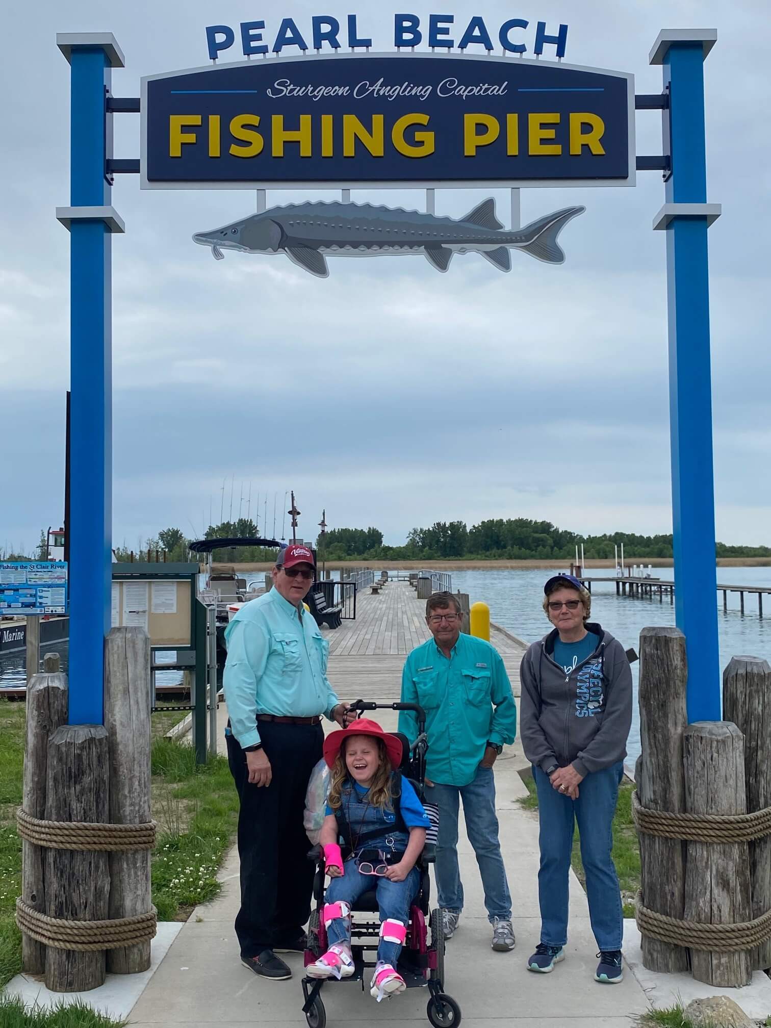 Group of people at Pearl Beach Fishing Pier. A person in a wheelchair is in the center, a large sign overhead depicts a fish.