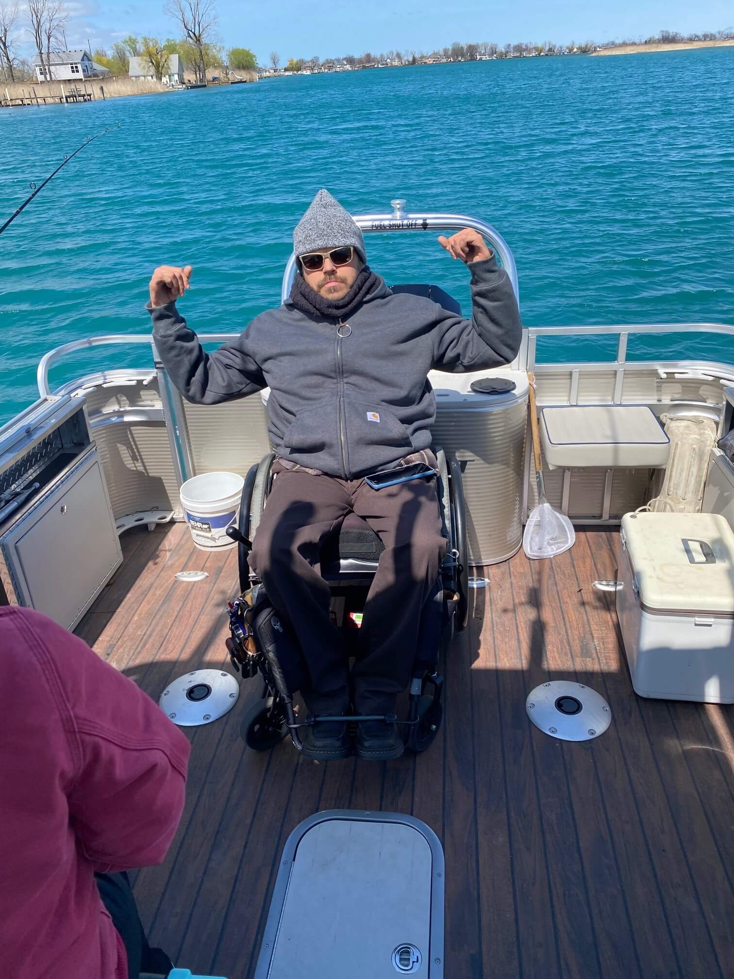 Man in wheelchair on a boat, flexing arms. Wearing a gray hat, jacket. Fishing on a bright blue lake.