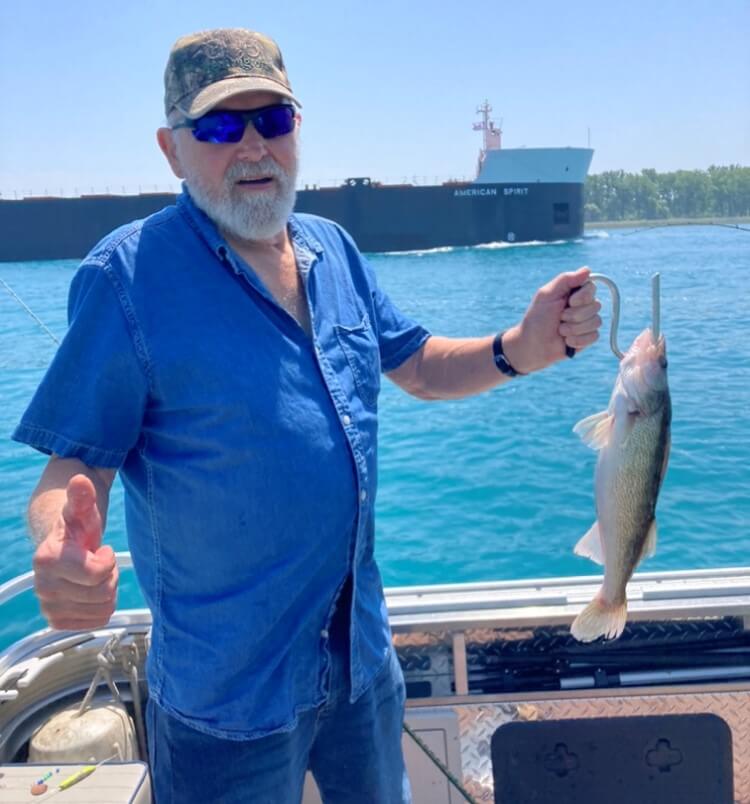Man on a boat, holding up a fish on a hook. He gives a thumbs-up. Blue sky, water, and a freighter visible.
