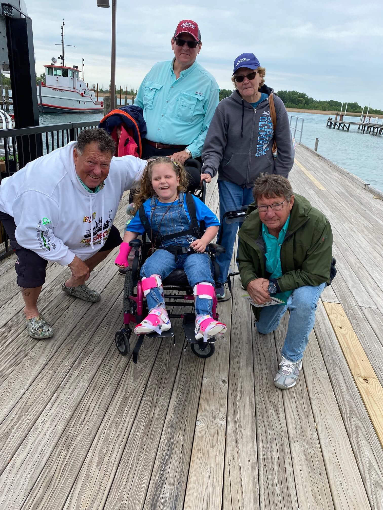 Group of people on a dock with a child in a wheelchair. One person kneels, others stand and smile, a boat is in the background.