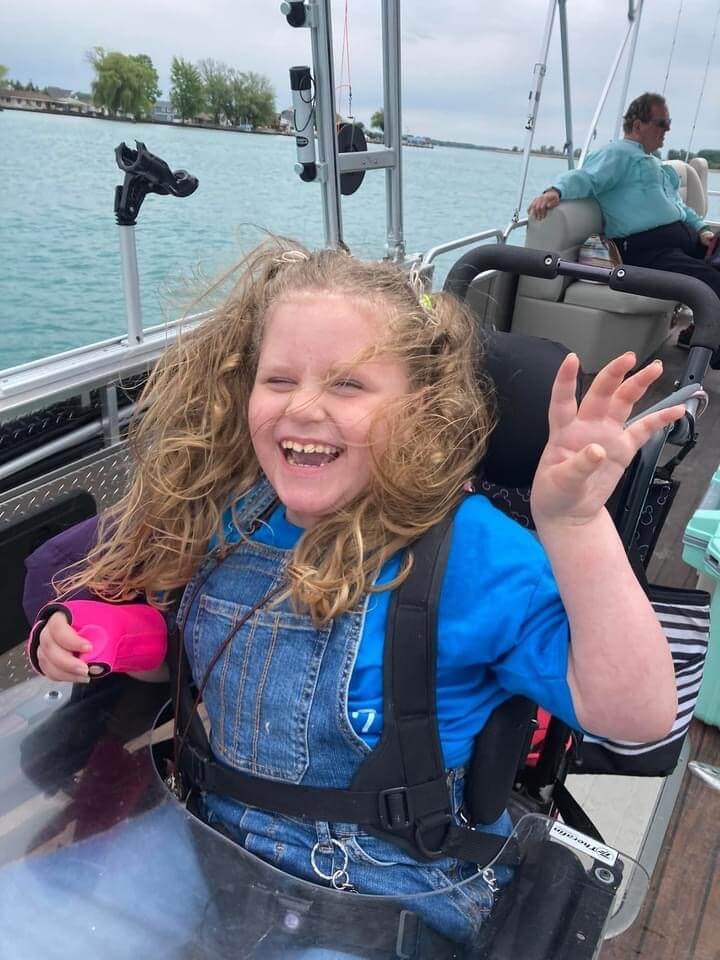 Smiling child in a wheelchair on a boat, waving, with water and another person visible in the background.