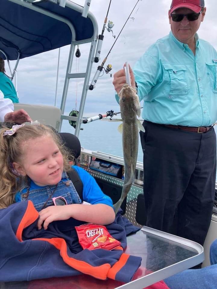 Man holds a fish on a boat; a child looks on. Overcast day; sea in the background.