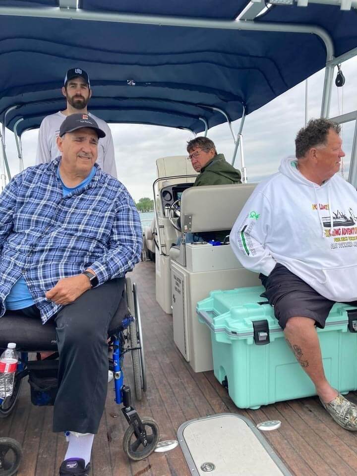 People on a pontoon boat, one in a wheelchair. Cloudy sky. Man in a wheelchair smiles. Others look ahead.