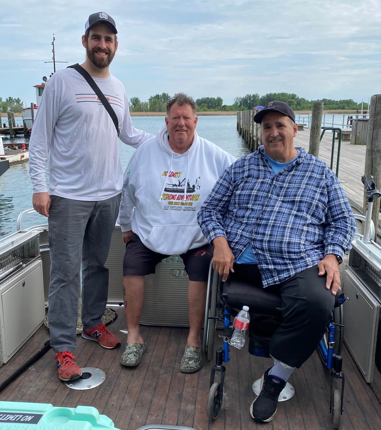 Three people on a boat. One in a wheelchair, two standing. Dock in background. Cloudy sky, water.