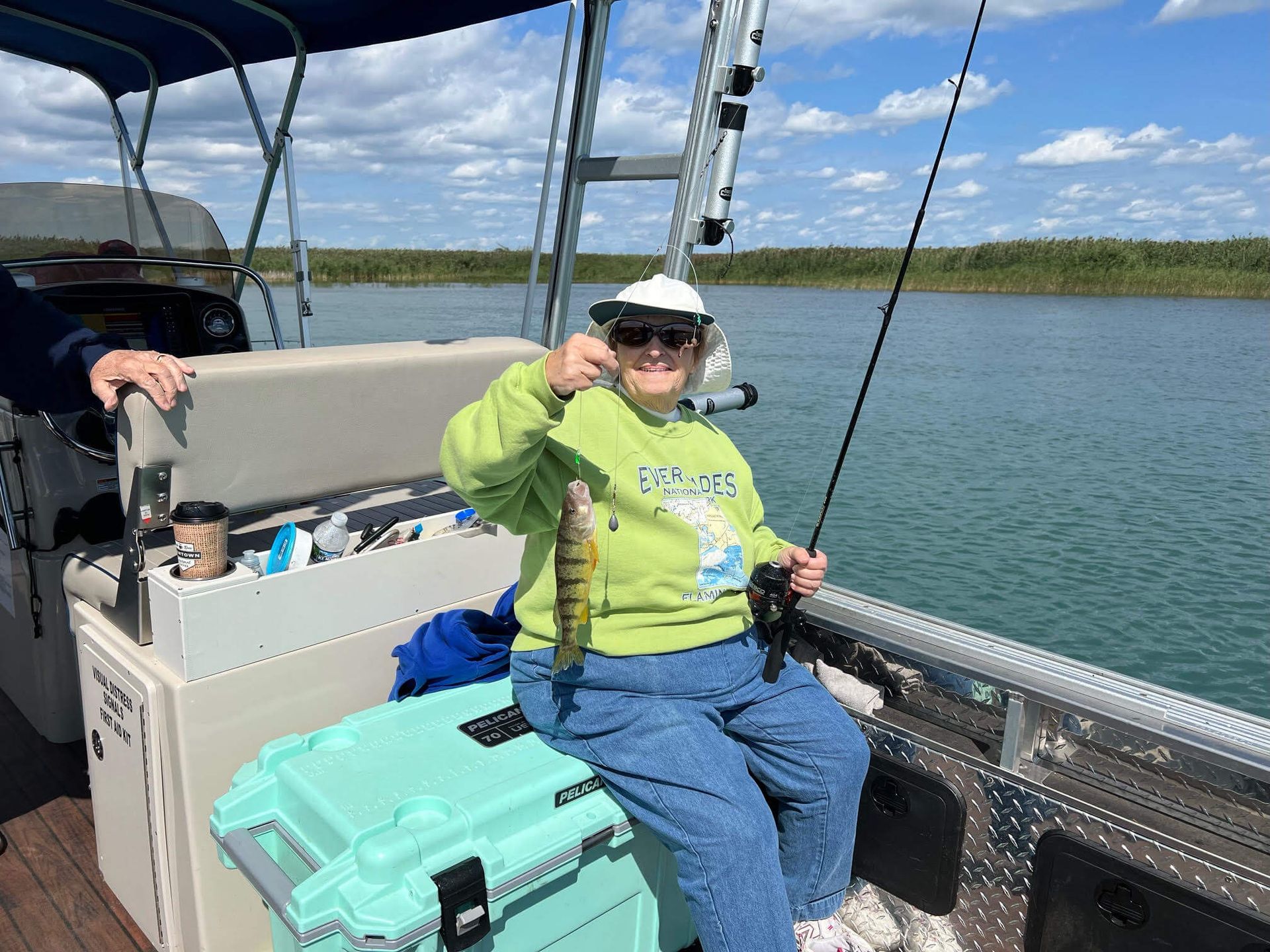 Woman on boat holds up a yellow perch, smiling. Blue water, sunny day.