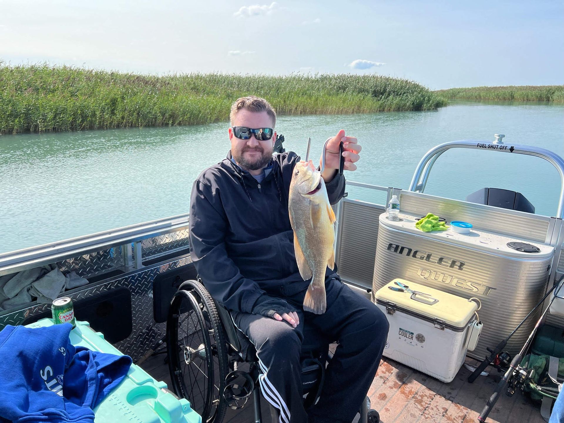 Man in wheelchair on boat, holding fish. Lake, reeds, and cooler in background.