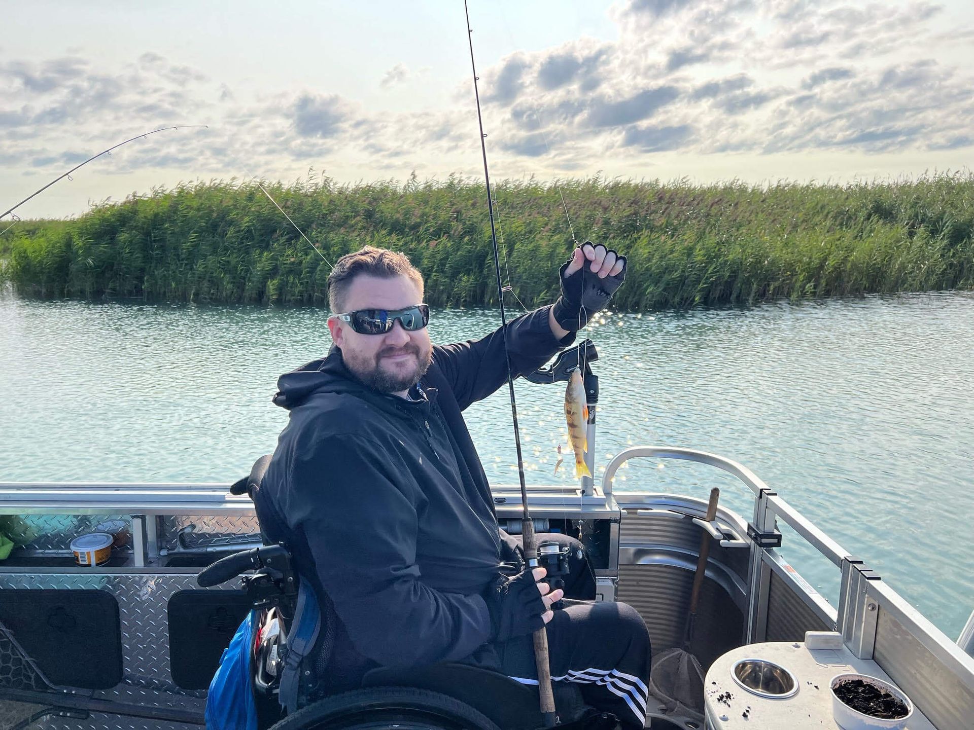 Man in a wheelchair fishing from a boat, holding up a small fish, with reeds in the background.