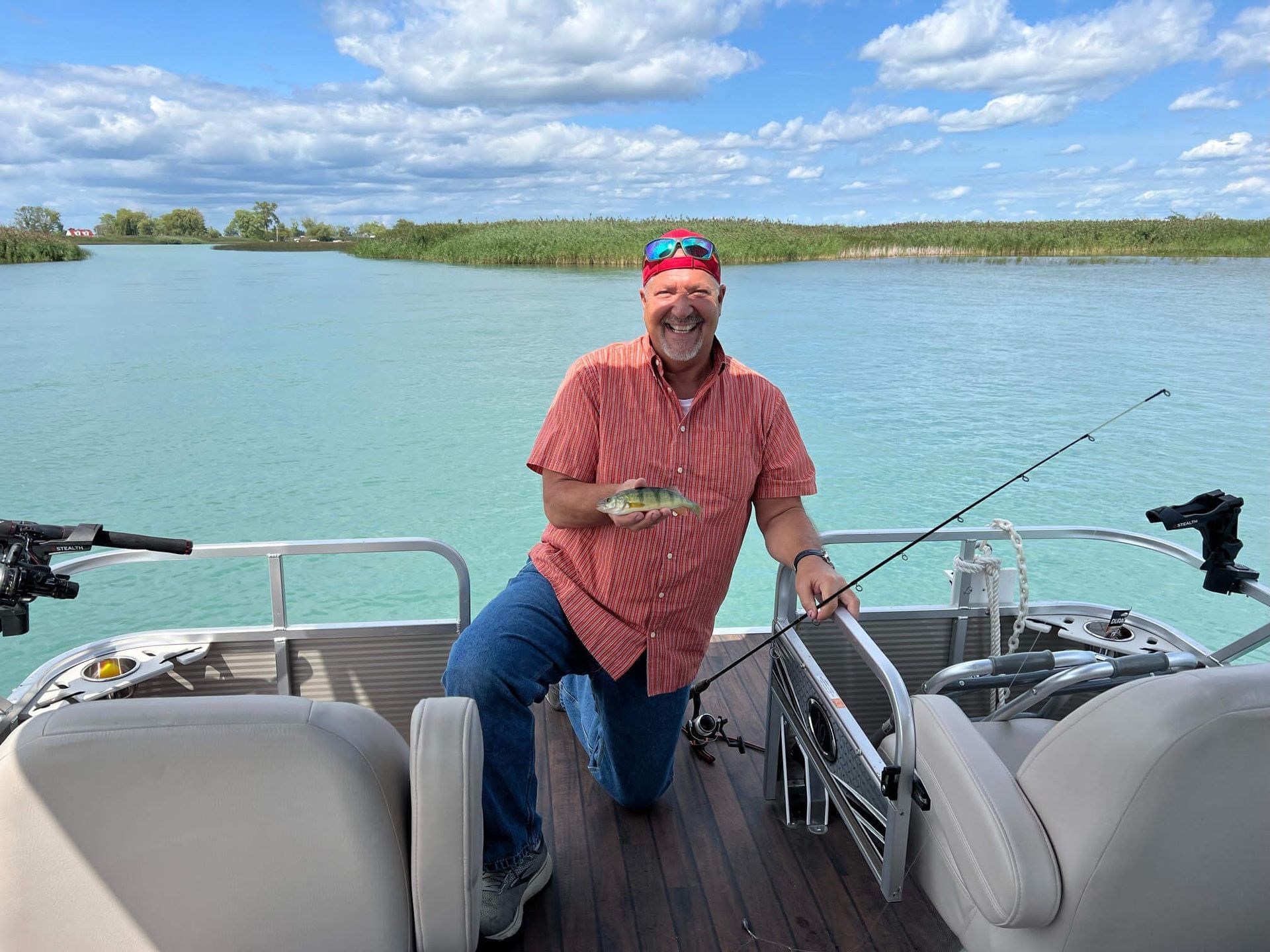Man on boat, holding fish. Wearing red shirt and bandana, fishing rod in hand. Turquoise water, sunny day.