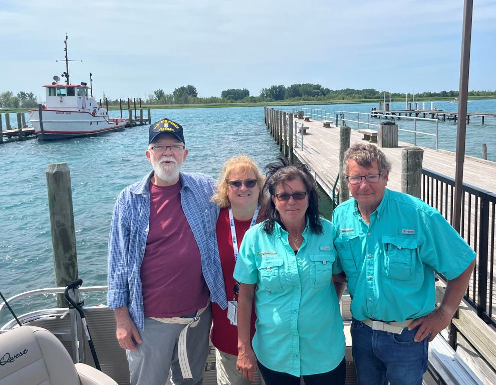 Four people pose on a dock with a boat and water in the background on a sunny day.