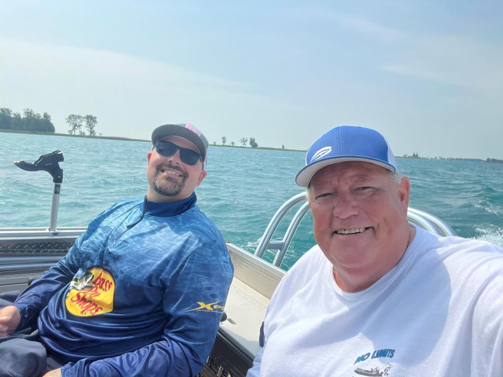 Two men on a boat, smiling, on a sunny lake. One wears sunglasses, the other a baseball cap.