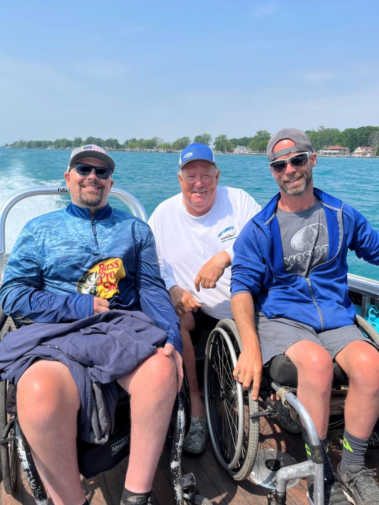 Three men in wheelchairs on a boat on a sunny day. They are smiling. Lake with houses in background.