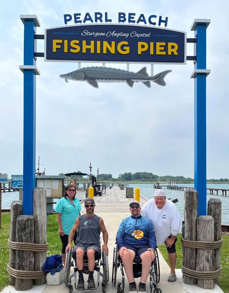 Four people by the Pearl Beach Fishing Pier sign. Two in wheelchairs, all smiling, the pier stretches to the water.