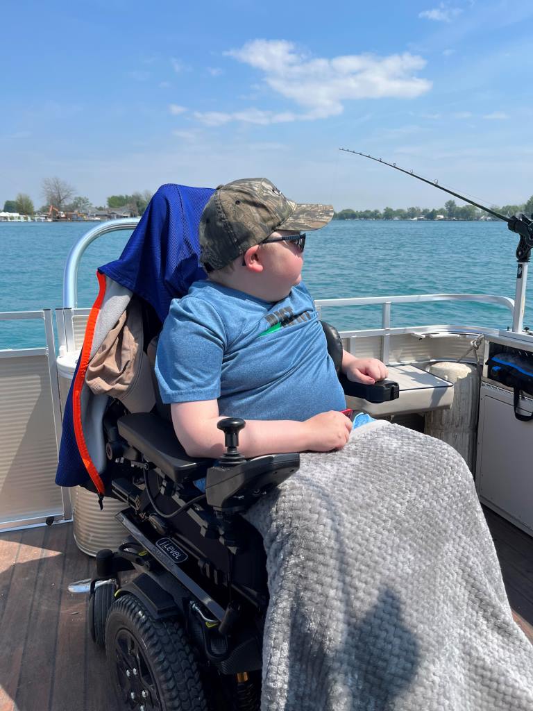 Person in wheelchair fishing on a boat, wearing hat and sunglasses, overlooking water.