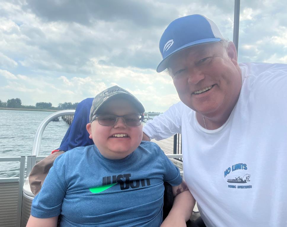 Two people on a boat smile at the camera. Cloudy sky, water background. The person on the right wears a white hat, white shirt.