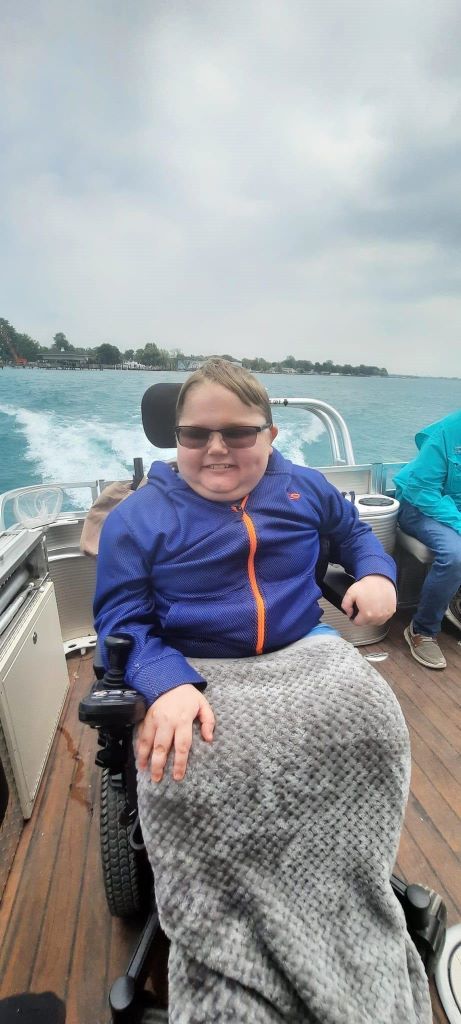 Person in a wheelchair smiles on a boat, wearing a blue jacket and sunglasses, with ocean and land in the background.