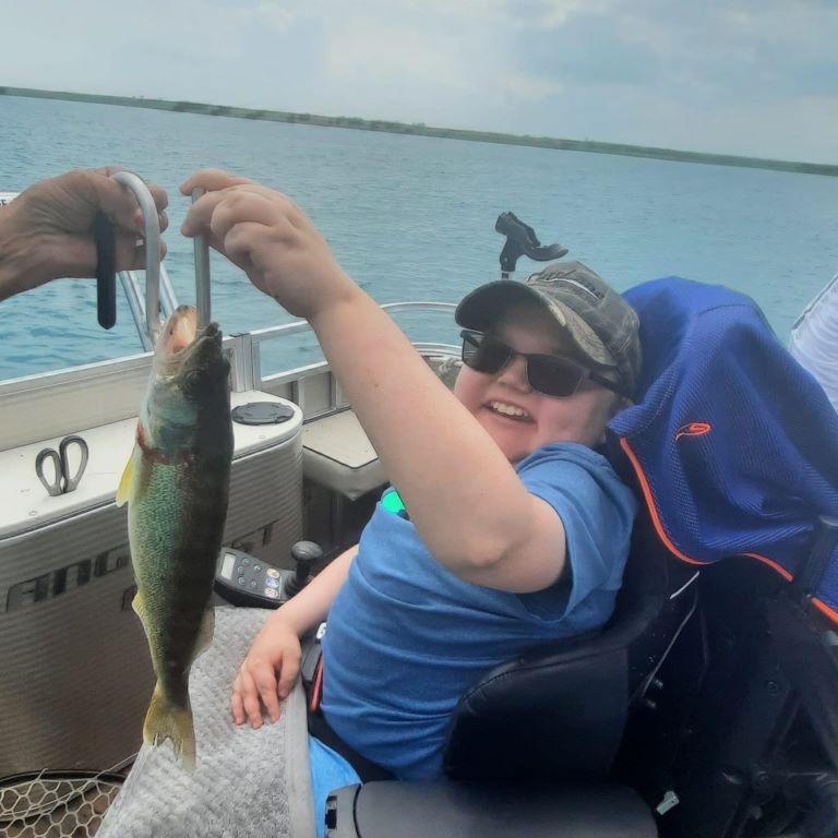 Person in wheelchair smiles, holding a fish on a boat. Another person assists with a scale; blue water in the background.